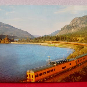 May include: A vintage postcard featuring a long orange passenger train with a glass-domed observation car traveling along a curved track beside a large blue river. In the background, green forested mountains rise under a clear blue sky.