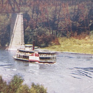 May include: A vintage steamboat with red and white accents sails on a river. The boat is passing under a bridge with a concrete support. Trees and foliage line the riverbanks, creating a scenic view.