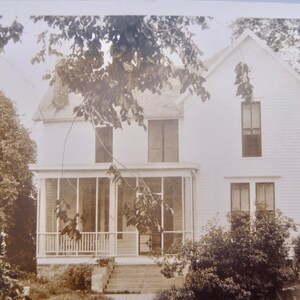 May include: A white two-story house with a screened-in porch. The house has a gabled roof and several windows. There are trees and bushes in the front yard.
