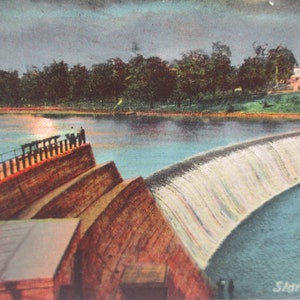 May include: A vintage postcard depicting a dam with a waterfall flowing over it. The dam is made of stone and has a wooden railing. There are people standing on the railing, looking out at the water. The water is a light blue color and the sky is a light gray color. The postcard says "Storage Dam" at the bottom.