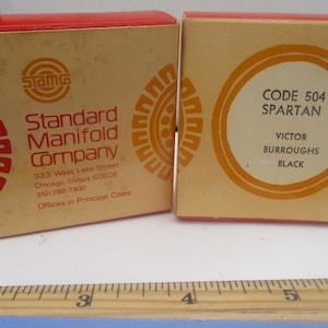 May include: Two vintage cardboard boxes with red and orange accents. One box features the text "Standard Manifold Company" and the other reads "CODE 504 SPARTAN". The boxes are tan with red and orange circular designs.