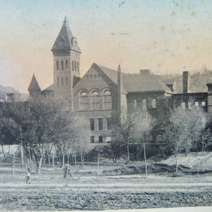 May include: A black and white vintage photograph of a brick building with a clock tower. The building has multiple windows and a large, open area in front of it.