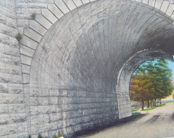 May include: A stone archway with a view of a tree-lined road leading to a body of water. The archway is made of large, rectangular stones.