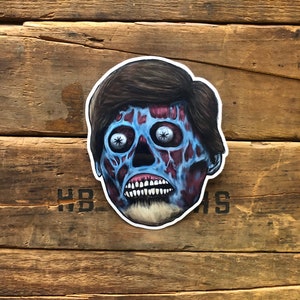 May include: A sticker featuring a zombie head with blue skin, exposed red tissue, and dark brown hair. The zombie has white eyes with star-shaped pupils and a wide, toothy grin. The sticker is set against a wooden background.