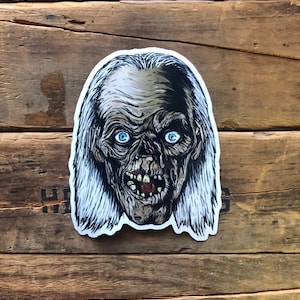 May include: A sticker of a zombie with long white hair and blue eyes. The zombie is grinning with its mouth open and teeth showing. The sticker is on a wooden background.