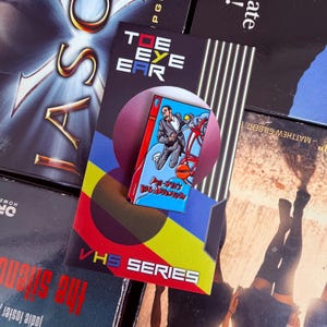 May include: A rectangular enamel pin featuring a Pee-wee's Big Adventure VHS cover design, displayed on a backing card labeled TOE EYE EAR and VHS SERIES. The pin depicts a character riding a red bicycle against a blue background.