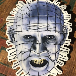 May include: A sticker of a white face with a black grid pattern and many silver pins sticking out. The face is contorted in a grimace with a wide open mouth and yellow teeth. The sticker has a white border and the text "@Toe_Eye_Ear" in the bottom right corner.