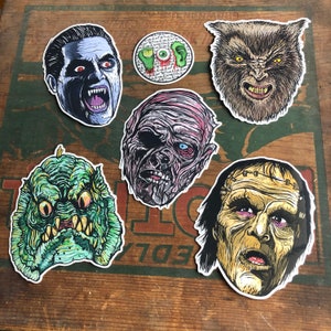 May include: A collection of horror-themed stickers featuring various monsters. The stickers include a vampire, a werewolf, a mummy, Frankenstein's monster, a creature from the black lagoon, and a sticker with an eye and ear design. The stickers are on a wooden surface.