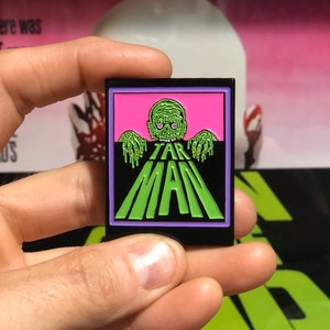 May include: A black enamel pin with a green and pink image of a zombie with the text "TAR MAN" in green letters.