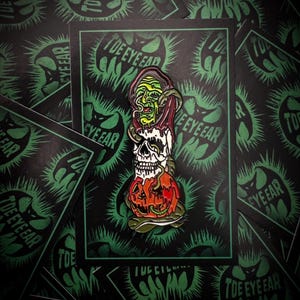 May include: A green, black, and orange enamel pin featuring a skull, a pumpkin, and a green snake wrapped around a woman's head. The pin is on a black background with green spikes.