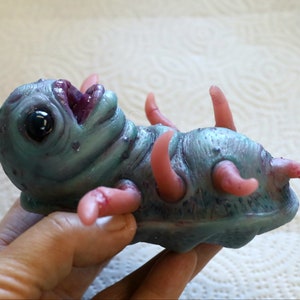 May include: A small, blue and purple creature with pink appendages. The creature has a large, open mouth and a single, black eye. It is made of a soft, rubbery material.