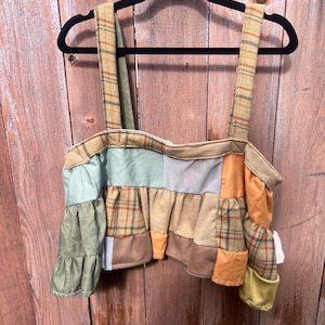 May include: A patchwork crop top with plaid and solid fabric panels in shades of green, tan, and orange. The top has wide straps and a ruffled hem. The garment is hanging on a black hanger against a wooden background.