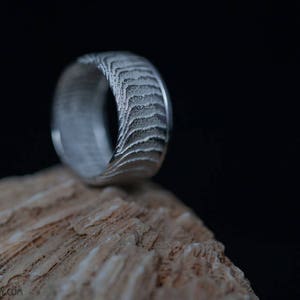 Puede incluir: Un anillo plateado con un patrón texturizado único, apoyado sobre una superficie rugosa de color marrón claro. La superficie del anillo presenta un diseño en capas, similar a olas. El fondo oscuro resalta los detalles del anillo.