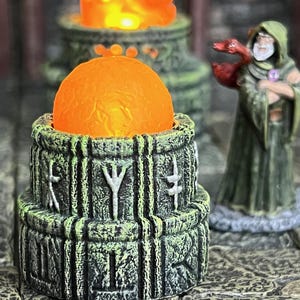 May include: A detailed image of a fantasy-themed miniature scene. A glowing orange orb sits atop a weathered, green and gray stone structure with rune carvings. A robed figure with a staff and a red dragon stands nearby.