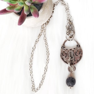 May include: A silver necklace with a heart-shaped padlock pendant and a black lava rock bead. The necklace is on a white background.