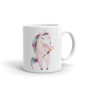 May include: A white ceramic mug featuring a pink unicorn holding a gold star wand. The unicorn has a rainbow mane and tail.