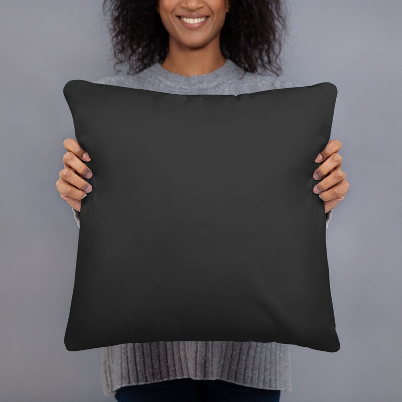 May include: A square black pillow held by a person. The pillow is a solid, dark color, and appears to be made of a soft fabric. The person is wearing a gray sweater.