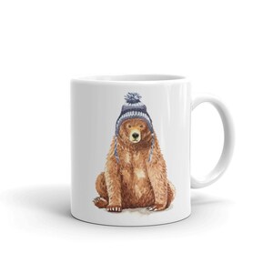 Peut inclure: Mug en céramique blanche avec une illustration à l'aquarelle d'un ours brun portant un bonnet en tricot bleu et gris avec un pompon. L'ours est assis et la tasse a une anse blanche.