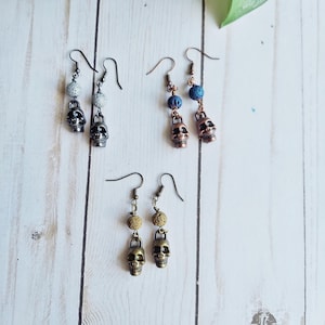 May include: Several pairs of skull earrings with different color beads. The earrings have a skull charm and are made of metal. The beads are gray, blue, and tan. The earrings are displayed on a white wooden surface.