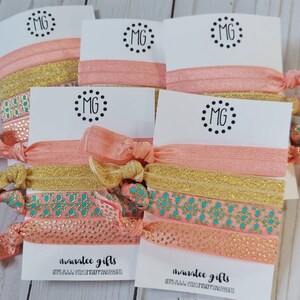 May include: Set of five hair scrunchies with a peach and gold color scheme. The scrunchies have a patterned design with turquoise and gold accents. The scrunchies are packaged on white cards with the text "manatee gifts" and a website address.