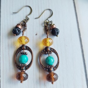 May include: A pair of handcrafted earrings featuring a combination of colors and textures. The earrings have copper-colored metalwork, with a turquoise bead, a yellow bead, and a brown faceted bead. The earrings also have a black lava bead.