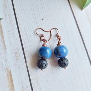 May include: A pair of handmade earrings featuring blue ceramic beads and dark gray lava stone beads. The earrings have copper-colored ear wires and connectors. The blue beads have a glossy finish and the lava beads have a textured surface.