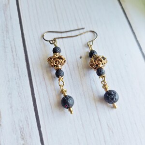 May include: A pair of dangle earrings featuring black lava beads and ornate gold-colored metal beads. The earrings have a long, slender design with a hook closure. The earrings are displayed on a light-colored wooden surface.
