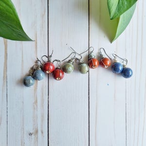 May include: A collection of handmade ceramic ball earrings in various colors, including blue, red, orange, and green. Each earring features a wire-wrapped design and a simple hook. The earrings are displayed on a white wooden surface.