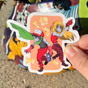 Puede incluir: Una pegatina con dos personajes de dibujos animados, un niño y una niña, con atuendos rojos y verdes. El niño sostiene una pistola azul y la niña sostiene una pistola roja. El fondo es de color melocotón con triángulos amarillos.