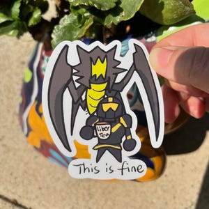 Può includere: Un adesivo nero e giallo con un cavaliere dei cartoni animati che tiene una tazza di tè. Il cavaliere indossa un'armatura e una corona in testa. L'adesivo dice "This is fine" e "Liber Tea" sulla tazza.