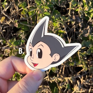May include: A sticker featuring the head of a cartoon character with black hair, large eyes, and a red mouth. The character's skin is light, and the sticker has a white border. The sticker is held against a background of green foliage.