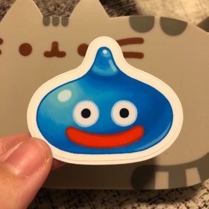 May include: A blue and red cartoon slime character sticker with a white border. The slime has two large eyes and a wide smile.