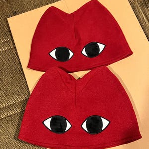 Puede incluir: Dos gorros de forro polar rojo, cada uno con un diseño de gato y grandes ojos negros y blancos. Los gorros tienen forma redondeada, probablemente para disfraz o uso de novedad.