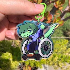 Puede incluir: Un llavero iridiscente verde y azul con un personaje de dibujos animados en una silla de ruedas. El personaje tiene el pelo verde, una cola verde y lleva una sudadera con capucha morada y zapatillas marrones.