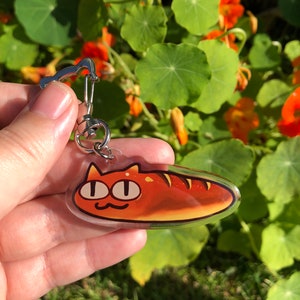 May include: A clear acrylic keychain featuring a cartoon cat shaped like a loaf of bread. The cat has orange fur, large eyes, and a small smile. The keychain is attached to a silver metal ring.