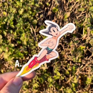 May include: A sticker featuring a cartoon character in a flying pose. The character has black hair, a red and yellow rocket, and is wearing black shorts and red boots. The letter "A" is in the lower left corner.