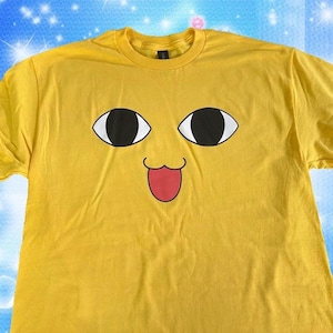 以下が含まれることがあります： 漫画の顔のデザインが施された黄色のTシャツ。顔には、大きな黒と白の目、赤い舌、笑顔の口があります。Tシャツは柔らかい素材でできており、半袖です。