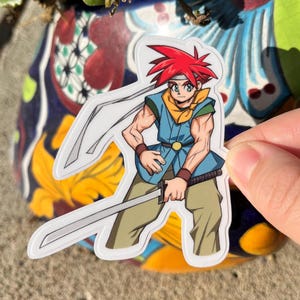 Peut inclure: Un autocollant représentant un personnage de dessin animé avec des cheveux roux, une chemise bleue et une ceinture jaune tenant une épée. Le personnage porte un bandeau blanc et a une expression déterminée.
