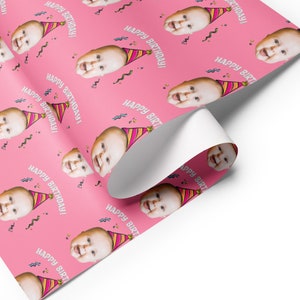 May include: Pink wrapping paper with a repeating pattern of a baby's face wearing a party hat and the text "Happy Birthday!"
