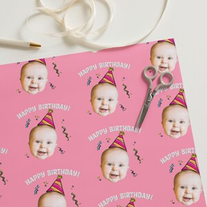 May include: Pink wrapping paper with a repeating pattern of a baby's face wearing a party hat and the text "Happy Birthday!"