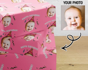 May include: Pink wrapping paper with a repeating pattern of a baby face wearing a party hat and the text "Happy Birthday!"