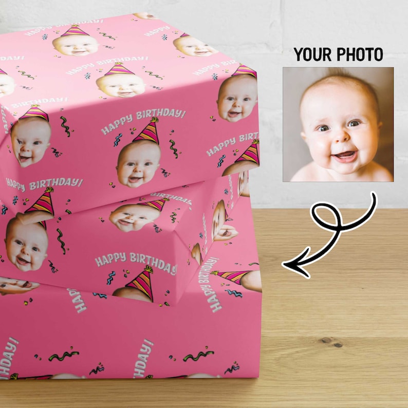 May include: Pink wrapping paper with a repeating pattern of a baby face wearing a party hat and the text "Happy Birthday!"
