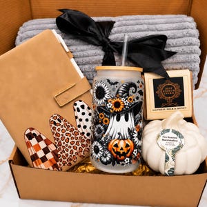 May include: A gift box containing a tan notebook with ghost illustrations, a frosted glass tumbler with a ghost and pumpkin design, a grey ribbed blanket with a black ribbon, a rectangular soap bar, and a white pumpkin-shaped candle.