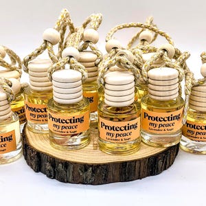 May include: A group of small glass bottles with cork stoppers and twine hangers. The bottles are filled with a clear liquid and have labels that say "Protecting my peace Lavender & Sage". The bottles are arranged on a wooden slice.