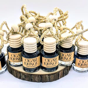 May include: A group of 12 black glass car air fresheners with wooden tops and gold string hangers. Each bottle has a label that reads "CIGAR LOUNGE Tobacco & Vanilla".