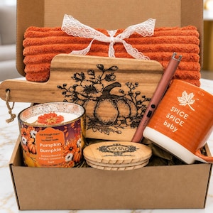 May include: A gift box containing an orange ribbed towel with a white lace ribbon, a wooden cutting board with a black pumpkin and floral engraving, a candle labeled Pumpkin Bumpkin, a wooden lid, and an orange mug with the text Spice Spice baby.