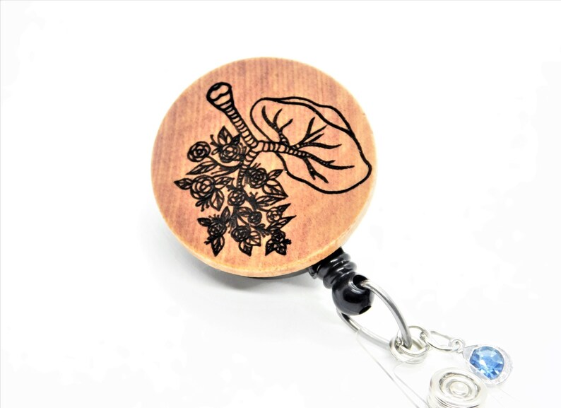 May include: A wooden badge reel with a black retractable cord and a blue crystal charm. The wooden circle features a black and white laser engraved design of a stylized lung with flowers.