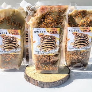 May include: Several clear pouches of scented wax, labeled "Sweet Tooth" with an image of pancakes. The pouches are filled with a light brown wax speckled with golden granules. Each pouch contains 7.5 ounces of Maple Pecan Pancakes squeeze wax.
