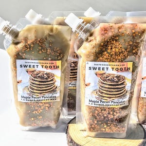 May include: Several clear plastic pouches filled with a light brown wax, speckled with brown and gold granules. Each pouch has a white spout and a label that reads "SWEET TOOTH" and "Maple Pecan Pancakes 7.5 OUNCE SQUEEZE WAX".