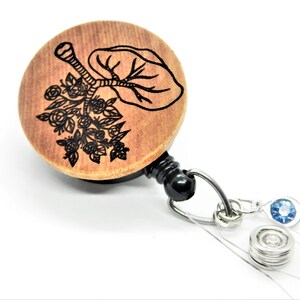 May include: Wooden badge reel with a black and white engraved design of a tree with flowers. The reel has a black retractable cord and a silver clip. The reel is attached to a clear plastic strap with a silver metal ring and a blue rhinestone.